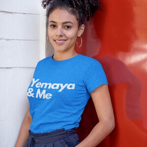 Puede incluir: Una mujer con una camiseta azul con el texto "Yemaya & Me" en letras blancas. Está de pie frente a una pared roja.