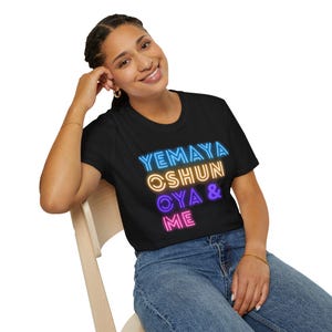 Può includere: Una maglietta nera con il testo "YEMAYA OSHUN OYA & ME" in colori al neon. Il testo è impilato verticalmente e i colori sono blu, giallo, viola e rosa.