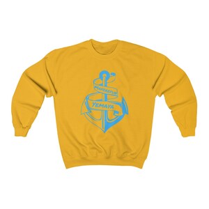 Puede incluir: Una sudadera amarilla con un gr&aacute;fico de ancla azul. El ancla est&aacute; envuelta con una pancarta que dice "MARESFUL YEMAYA".