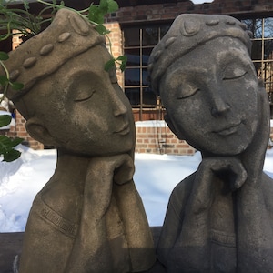 May include: Two gray stone sculptures of human heads with closed eyes, each with a crown on top. The sculpture on the left is a planter with a green plant growing out of the top.