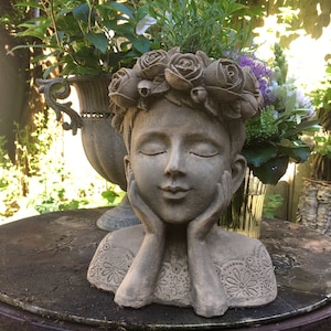 Puede incluir: Un macetero de piedra gris con forma de cabeza de mujer con una corona de flores. El macetero está sentado en una mesa de madera y tiene un diseño de encaje alrededor del cuello.