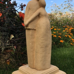 Könnte beinhalten: Eine beigefarbene Steinskulptur von zwei sich umarmenden Figuren, die auf einem mehrstufigen Sockel stehen. Die Skulptur befindet sich im Freien in einem Garten.