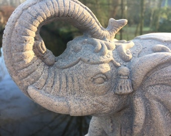Puede incluir: Una estatua de elefante de piedra gris detallada con tallas intrincadas y una trompa levantada. El ojo del elefante es visible y la estatua tiene una superficie texturizada.