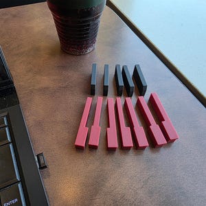 Könnte beinhalten: Eine Anordnung roter und schwarzer Klaviertasten-förmiger Objekte auf einer braunen Oberfläche. Die roten Tasten sind länger und breiter als die schwarzen Tasten. Eine Laptop-Tastatur und eine Tasse sind ebenfalls zu sehen.