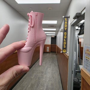 May include: A light pink platform boot with a high stiletto heel and a zipper closure. The boot has a lace-up design and is held up by a person's fingers. The boot is a miniature model.