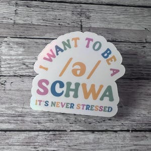 May include: A white sticker with the text "I WANT TO BE A /ə/ SCHWA" in various colors. Below the word "SCHWA" is the text "IT'S NEVER STRESSED". The sticker is on a wooden background.