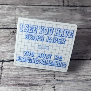 May include: A white square sticker with blue text that reads "I SEE YOU HAVE GRAPH PAPER... YOU MUST BE PLOTTING SOMETHING." The sticker has a grid pattern and is set against a wooden background.