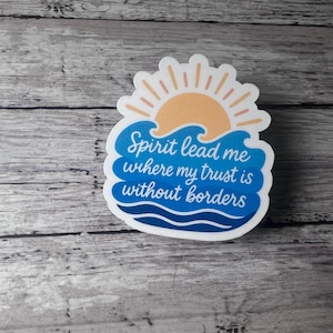 Puede incluir: Una pegatina con un diseño que presenta un sol amarillo, olas azules y el texto "Spirit lead me where my trust is without borders". La pegatina tiene un borde blanco y está sobre una superficie de madera.