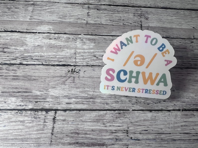 May include: A white sticker with the text "I WANT TO BE A /ə/ SCHWA" in various colors. Below the word "SCHWA" is the text "IT'S NEVER STRESSED". The sticker is on a wooden background.