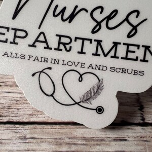 May include: White sticker with black text that reads "Nurses Department Alls Fair in Love and Scrubs". A black stethoscope is drawn in the shape of a heart with a feather inside.