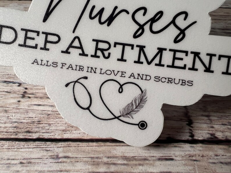 May include: White sticker with black text that reads "Nurses Department Alls Fair in Love and Scrubs". A black stethoscope is drawn in the shape of a heart with a feather inside.