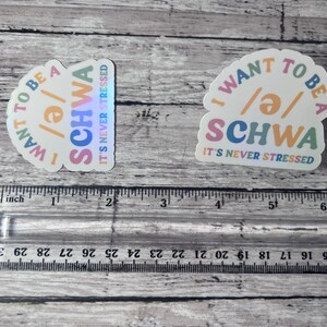 May include: Two stickers with the text "I WANT TO BE A /ə/ SCHWA IT'S NEVER STRESSED" in a colorful design. The stickers are on a wooden surface with a ruler in the foreground.