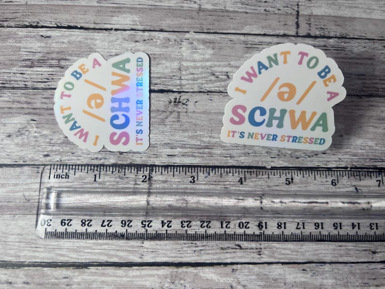 May include: Two stickers with the text "I WANT TO BE A /ə/ SCHWA IT'S NEVER STRESSED" in a colorful design. The stickers are on a wooden surface with a ruler in the foreground.