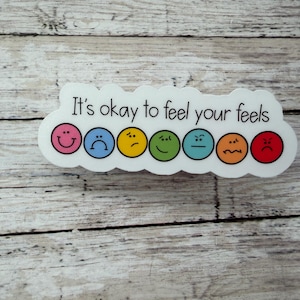 以下が含まれることがあります： It's okay to feel your feelsという黒い文字が書かれた白いダイカットステッカー。その下には、ピンク、青、黄色、緑、ターコイズ、オレンジ、赤の7つのカラフルな円形の顔が横一列に並んでおり、それぞれ異なる感情を表現しています。