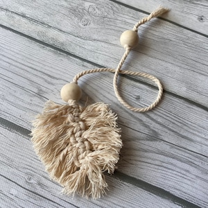May include: A beige macrame leaf with fringe hanging from a beige cord with two wooden beads.