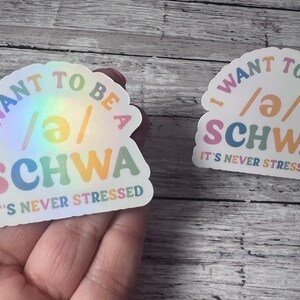 May include: Two holographic stickers with the text "I WANT TO BE A SCHWA IT'S NEVER STRESSED". The stickers have a rainbow sheen and feature colorful text in a playful font. The stickers are on a wooden surface.