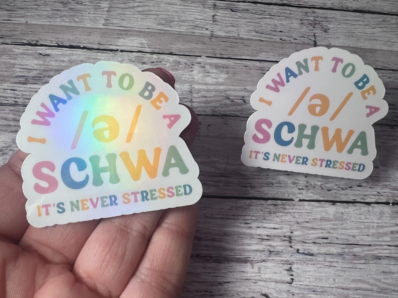 May include: Two holographic stickers with the text "I WANT TO BE A SCHWA IT'S NEVER STRESSED". The stickers have a rainbow sheen and feature colorful text in a playful font. The stickers are on a wooden surface.