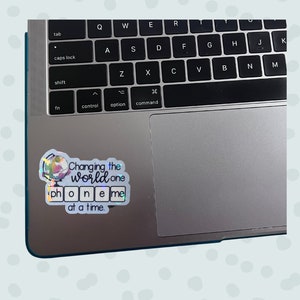 May include: A holographic sticker with a globe illustration and the text "Changing the world one phone me at a time." The sticker is placed on a silver laptop.