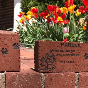 May include: Two red brick pavers with engraved messages honoring pets named Maxwell and Harley. The bricks are placed on a brick wall with a flower bed in the background.