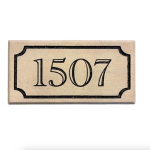 May include: A rectangular beige house number plaque featuring the black engraved digits 1507. The plaque has a decorative black border with rounded corners, creating a framed effect around the centered numbers on the light-colored surface.