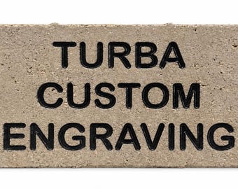 May include: A rectangular, light gray concrete brick featuring the black engraved text "TURBA CUSTOM ENGRAVING" centered on its front surface. The brick has a rough, porous texture and sharp, defined edges, presenting a clear, bold display of the lettering.