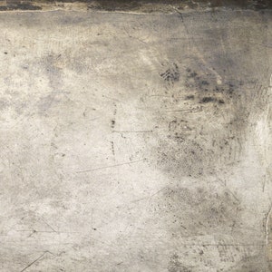 May include: A close-up of a textured, metallic surface with scratches and marks. The surface is a light gray color with darker gray streaks and spots.