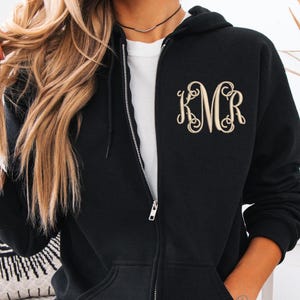 Peut inclure: Sweat à capuche noir zippé avec un monogramme doré sur la poitrine gauche. Le sweat à capuche a une capuche à cordon, une fermeture éclair sur toute la longueur et deux poches avant. Le texte "Custom Threads" est visible sur la manche droite.