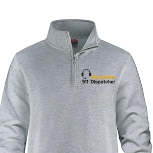 May include: A light grey sweatshirt with a quarter-zip collar. The text "Samantha 911 Dispatcher" is printed in yellow and black, with a headset graphic. The sweatshirt has long sleeves and a ribbed hem.
