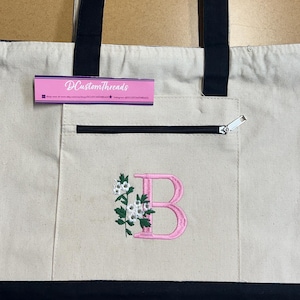 Peut inclure: Un sac fourre-tout en toile couleur crème avec des anses et une base bleu marine. L'avant présente une poche zippée, une lettre B rose brodée et un motif floral. Une étiquette violette avec le texte DCustomThreads est attachée.