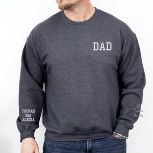 Peut inclure: Un sweat-shirt gris foncé avec le mot "DAD" brodé sur le devant. Les manches sont brodées des noms "THOMAS", "MIA" et "ALEXIA".