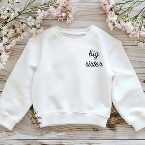 May include: A white long-sleeve crewneck sweatshirt featuring the black text "big sister" printed on the upper right chest area. The garment has ribbed cuffs and a ribbed hem, with a small "D Custom Threads" logo near the bottom left hem.