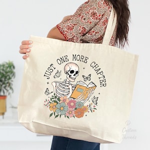 Peut inclure: Un sac fourre-tout en toile blanche avec le texte "Just One More Chapter" imprimé dessus. Le texte est entouré d'un motif floral et d'un squelette tenant un livre.