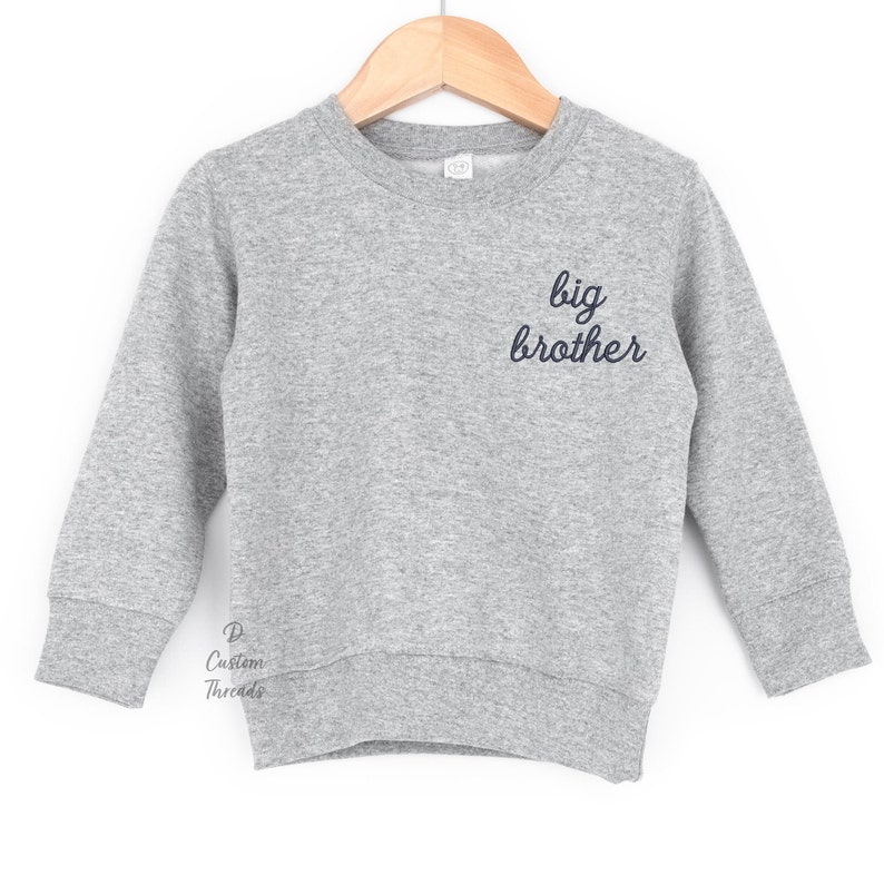 May include: A heather gray long-sleeve crewneck sweatshirt featuring the text "big brother" embroidered in dark navy blue script on the upper right chest. The garment is displayed on a wooden hanger and includes a small "D Custom Threads" logo.