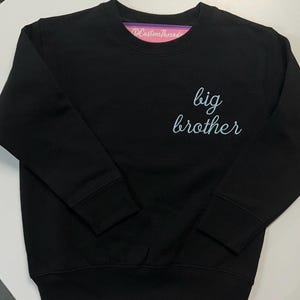 May include: A black long-sleeve crewneck sweatshirt featuring the text "big brother" in a light blue cursive script on the upper right chest. The garment has a ribbed collar, cuffs, and hem, with a pink brand label visible inside the neckline.