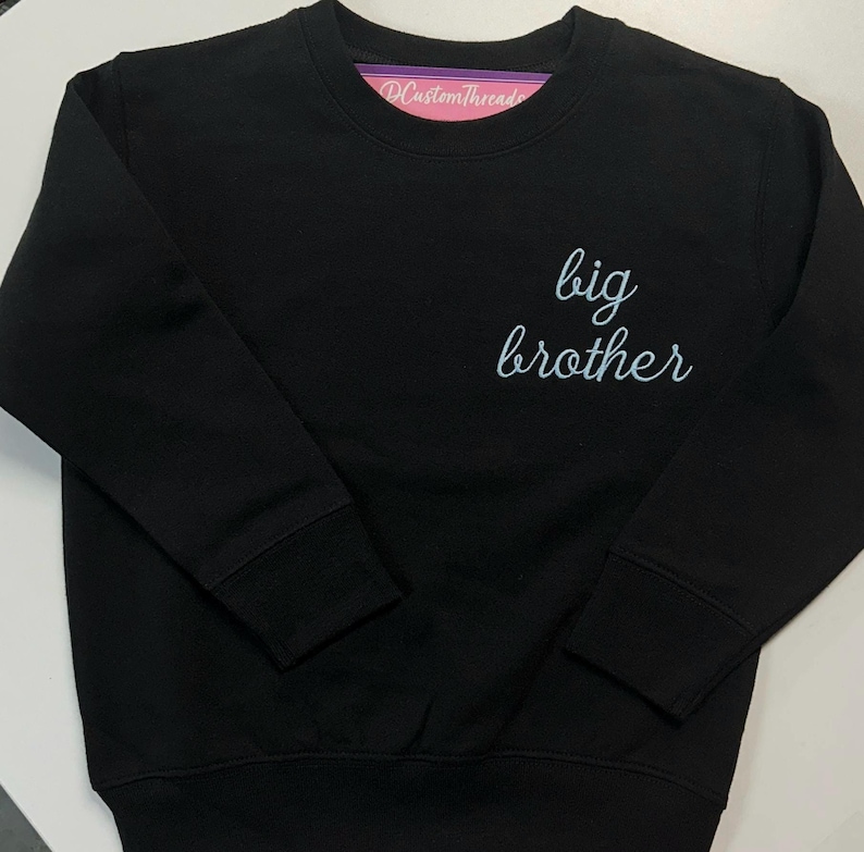 May include: A black long-sleeve crewneck sweatshirt featuring the text "big brother" in a light blue cursive script on the upper right chest. The garment has a ribbed collar, cuffs, and hem, with a pink brand label visible inside the neckline.