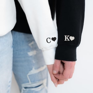 May include: Two people holding hands, wearing white and black sweatshirts with the letters "C" and "K" and a heart on each sleeve.