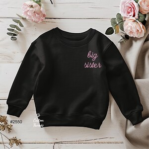May include: A black long-sleeve sweatshirt featuring the text big sister in a light pink cursive font on the upper left chest area. The garment has a crew neckline and ribbed cuffs at the sleeves and hem, with the text #2550 and Custom Threads visible.