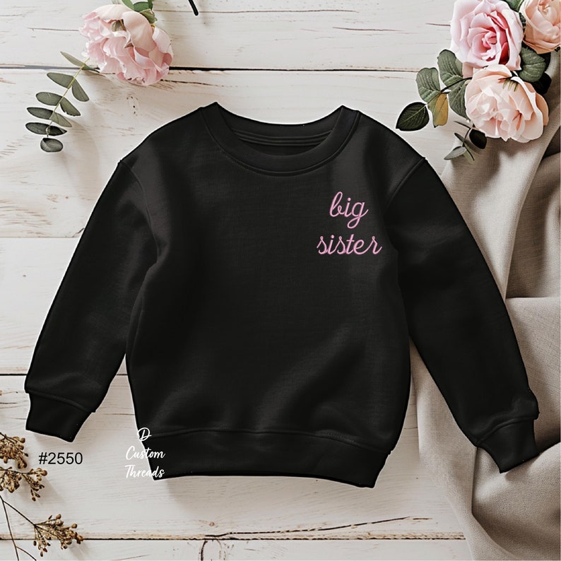 May include: A black long-sleeve sweatshirt featuring the text big sister in a light pink cursive font on the upper left chest area. The garment has a crew neckline and ribbed cuffs at the sleeves and hem, with the text #2550 and Custom Threads visible.