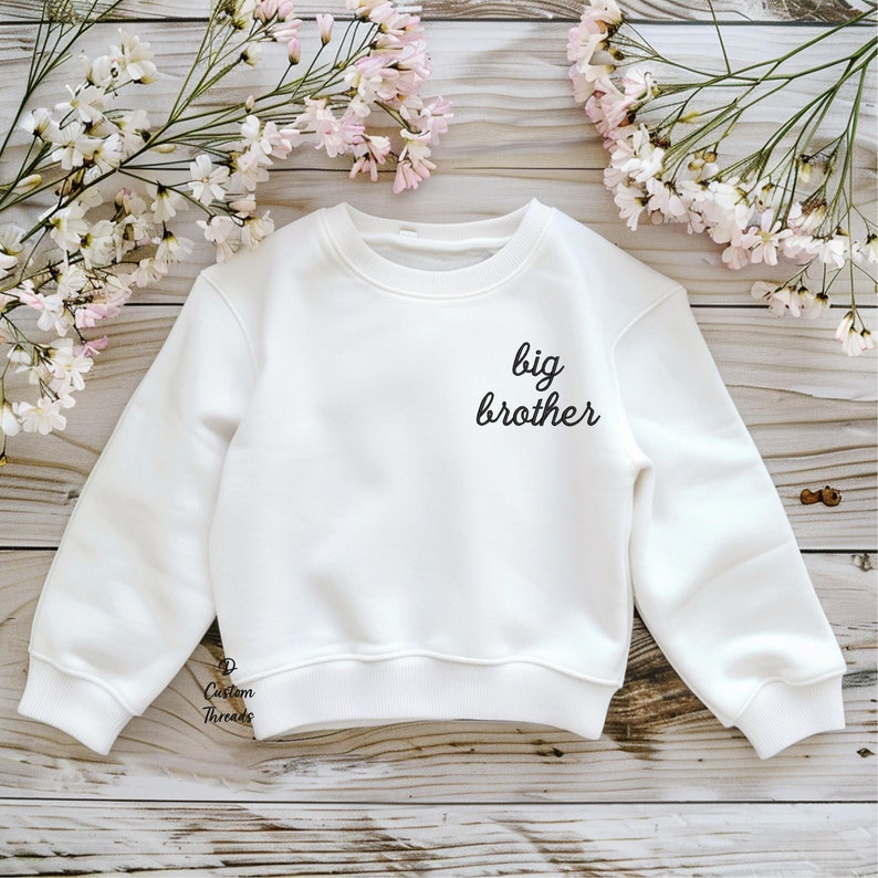 May include: A white long-sleeved crewneck sweatshirt featuring the black text "big brother" printed on the upper left chest area. The garment has ribbed cuffs and a ribbed hem, with a small "Custom Threads" logo printed near the bottom left.