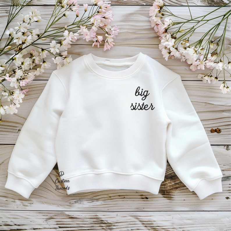 May include: A white long-sleeve crewneck sweatshirt featuring the black text "big sister" printed on the upper right chest area. The garment has ribbed cuffs and a ribbed hem, with a small "D Custom Threads" logo near the bottom left hem.