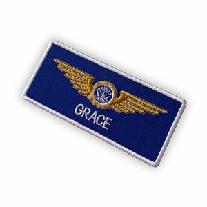 May include: A navy blue embroidered patch with a white border. It features gold-coloured wings and a central emblem. The word "GRACE" is embroidered in white below the emblem. This patch is likely a custom name tag or uniform accessory.