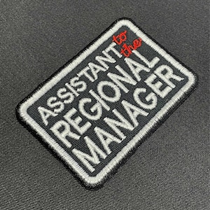 May include: A black and white embroidered patch with the text "Assistant to the Regional Manager" in white lettering. The word "to" is in red.