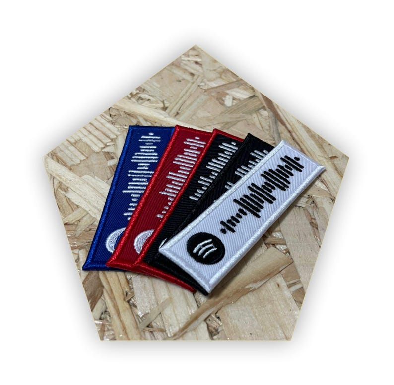 Op de afbeelding: Vijf geborduurde patches met een zwart-witte barcode-ontwerp. De patches zijn in blauw, rood, zwart en wit. De witte patch heeft een kleine witte cirkel met drie zwarte lijnen in het midden.