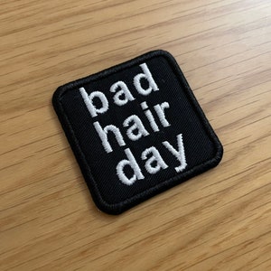 Może przedstawiać: Czarna łatka z tkaniny z białym haftem, na której widnieje napis "bad hair day".