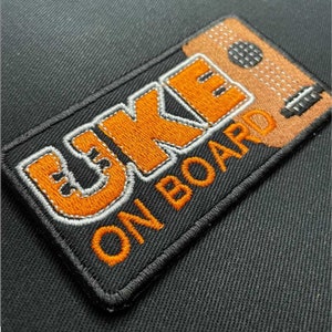 Może przedstawiać: Naszywka haftowana pomarańczowa i biała z tekstem "UKE ON BOARD".