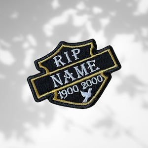 Könnte beinhalten: Schwarz-goldener, gestickter Aufnäher mit dem Text "RIP NAME 1900 2000" und einer weißen Taube. Der Aufnäher hat eine Schildform mit einem Banner in der Mitte. Die Texte "RIP" und "NAME" sind in Weiß.