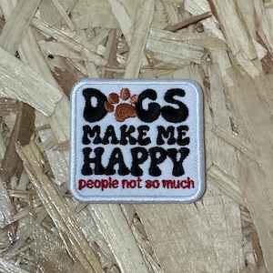 Può includere: Toppa ricamata bianca con testo nero che recita "I cani mi rendono felice, le persone non così tanto". Il testo è in uno stile di font retrò. C'è un'impronta di zampa marrone sopra la parola "Cani".