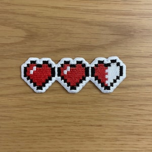 May include: Three pixelated hearts, two full red hearts and one half red and half white heart, embroidered on a white fabric patch.