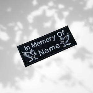 Könnte beinhalten: Schwarzer, rechteckiger Aufnäher mit weißer Stickerei. Der Text "In Memory Of Name" ist mittig platziert, flankiert von zwei stilisierten Adlerdesigns. Der Aufnäher dient wahrscheinlich Gedenkzwecken.
