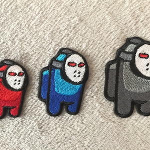 Op de afbeelding: Drie geborduurde strijkpatches met het Among Us-personage dat een hockeymasker draagt. De patches zijn rood, blauw en grijs.